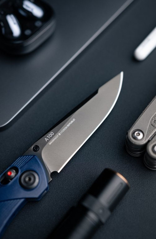 A100 Blue Magnacut ANV Lock Knife shown on a dark table as a closeup of the blade. Laying next to a torch and a multitool in typical EDC flatlay style. 